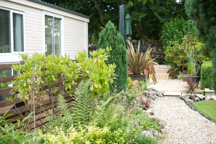 A modern grey and white static caravan with a wooden sun deck, parked on a manicured grass plot with woodland in the background.